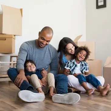 A family of four sits on the floor of their new home, surrounded by unpacked boxes and a warm, welcoming atmosphere.
