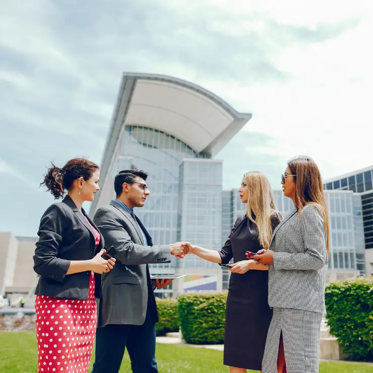 second three banner A group of four professionals shake hands and exchange documents outdoors, with a modern building in the background under a bright sky.